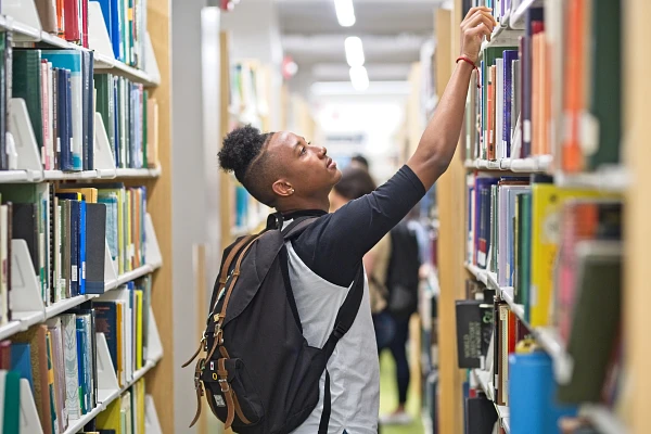 Student in a library reaches for book on a high shelf